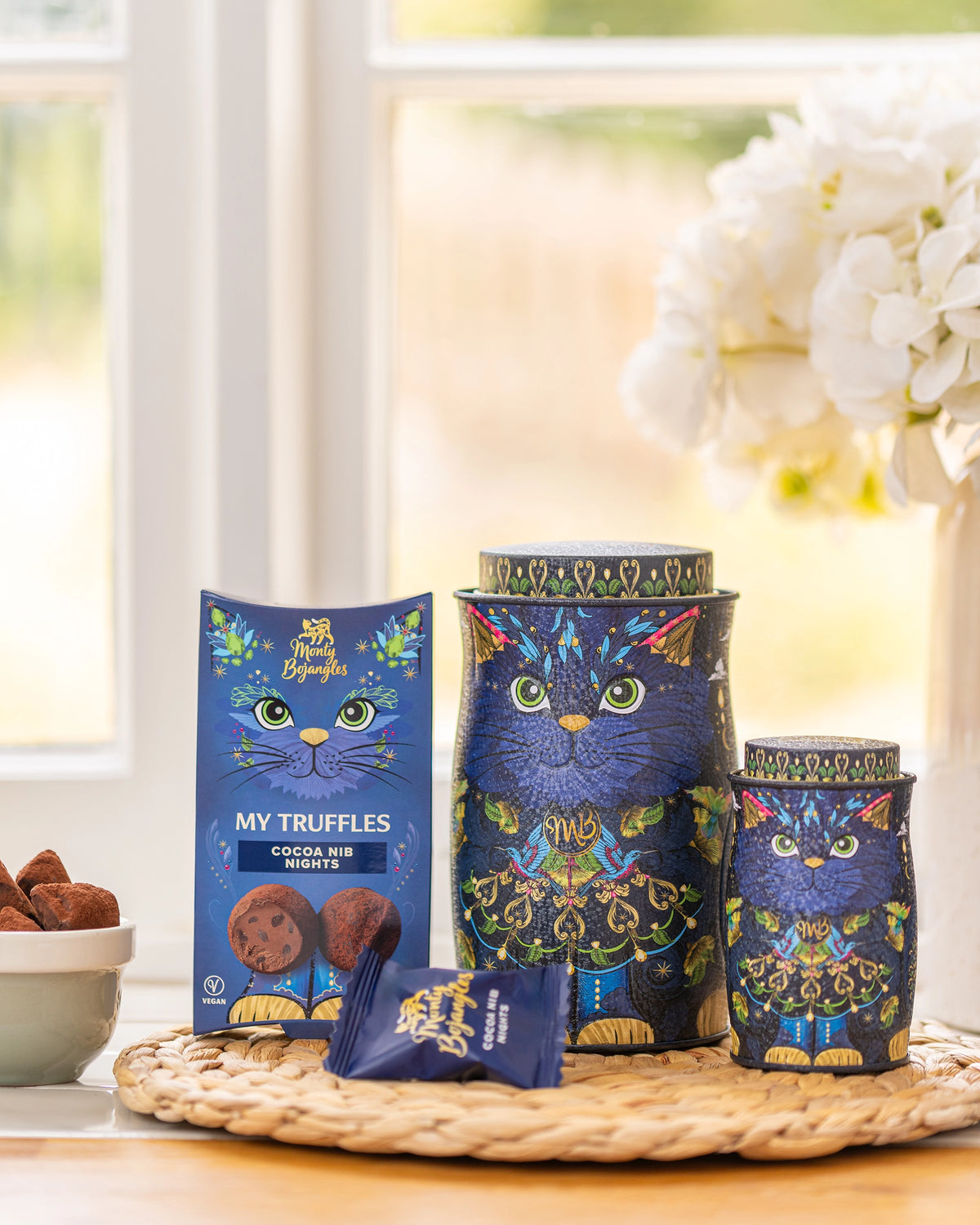Three blue cat-themed tin containers and a matching box of My Truffles chocolate truffles arranged on a woven tray in a bright kitchen setting.