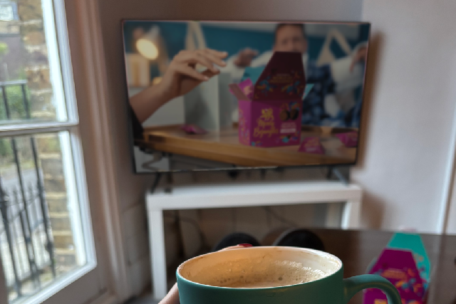 Person holding a mug with 'Monty Bojangles' branding, sitting on a sofa with a TV in the background.